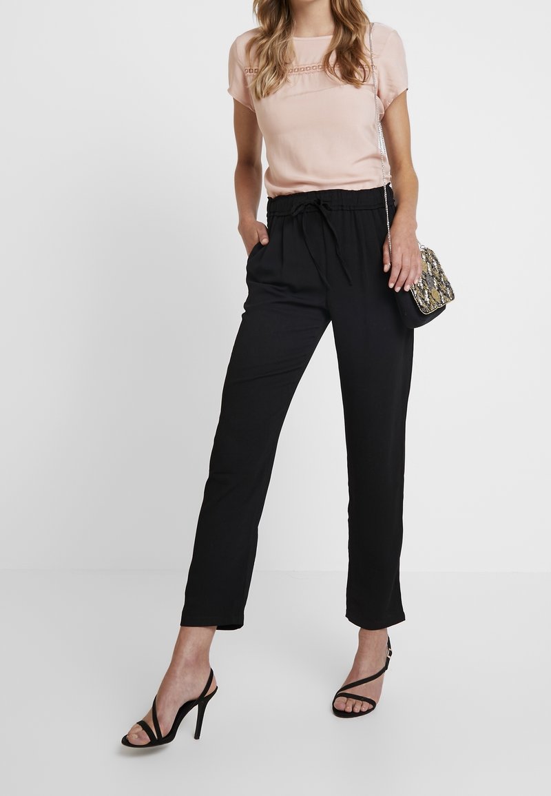 Czarne spodnie z elastycznym pasem i kieszeniami, połączone z jasnoróżową bluzką z ozdobnym przeszyciem. Otwarty czarny obcas.