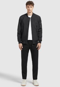 Schwarze Bomberjacke mit Reißverschlusstaschen, gerippten Bündchen und einem Frontreißverschluss, kombiniert mit schwarzen Hosen und hellen Sneakers.