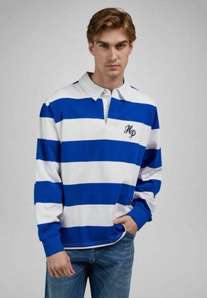 Jeune homme portant un polo à manches longues rayé bleu et blanc avec les initiales "HP" sur la poitrine et un jean bleu, posant devant un fond uni.