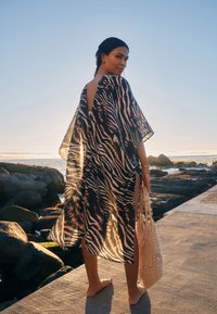 Schwarz-beiger Kaftan mit Zebra-Muster, offenem Rückenausschnitt, lockerem Sitz und seitlichen Schlitzen, kombiniert mit einer gewebten beigen Tragetasche. Sonnendurchfluteter Strandhintergrund.