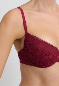 Primer plano de una mujer que lleva un sujetador de encaje burdeos con patrones florales y tirantes finos ajustables sobre un fondo claro.