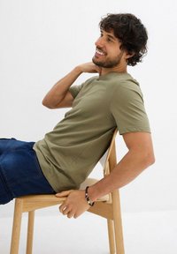 T-shirt verde oliva a maniche corte con collo rotondo. Il tessuto è liscio e la persona è seduta su una sedia di legno chiaro. Indossa jeans in denim scuro.