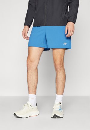 Piernas masculinas que llevan pantalones cortos deportivos azules de New Balance, calcetines blancos y zapatillas de correr blancas de New Balance, de pie sobre un suelo gris liso.