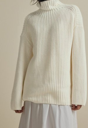 Personne portant un pull à col roulé en maille épaisse de couleur crème, à manches longues et coupe ample, par-dessus une jupe blanche.
