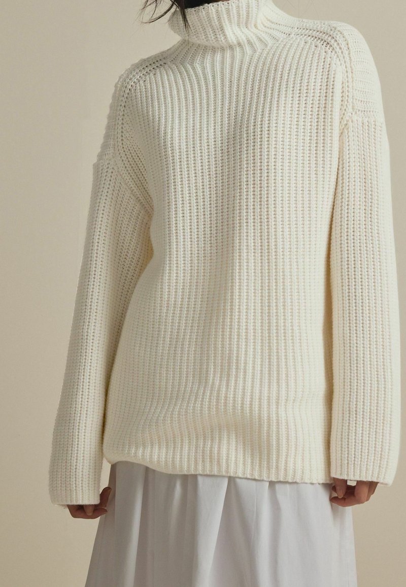 Personne portant un pull à col roulé en maille épaisse de couleur crème, à manches longues et coupe ample, par-dessus une jupe blanche.
