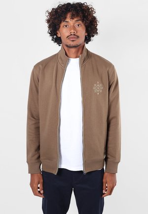 Homme aux cheveux bouclés portant une veste marron zippée par-dessus une chemise blanche et un pantalon sombre, se tenant devant un fond clair uni.