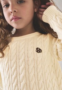 Šviesiai geltonas megztinis su pynimo raštu, apvalia apykakle ir ruda logotipo detalė. Riboti rankogaliai suteikia tekstūros ir dizaino.