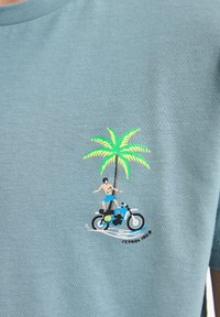 Camiseta de algodón azul claro con un gráfico bordado colorido de un hombre en motocicleta, una palmera, olas estilizadas y texto.