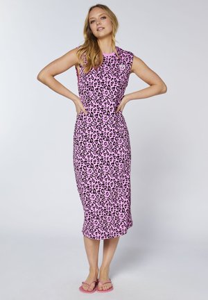 Frau trägt ein ärmelloses pinkfarbenes Midikleid mit schwarzem Leopardenmuster, steht mit den Händen in die Hüften gestützt und trägt pinke Flip-Flops.