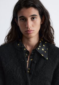 Schwarzer flauschiger Cardigan über einem schwarzen Hemd mit gelben Blumenmustern. Verfügt über einen Knopfkragen und eine strukturierte Oberfläche.