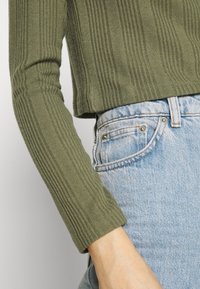 Top de manga larga en verde oliva acanalado, de forma ajustada, combinado con jeans de mezclilla azul claro que presentan detalles metálicos y un diseño típico de cinco bolsillos.