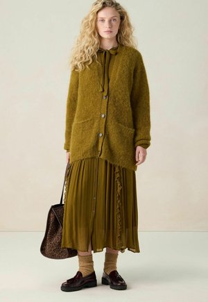 Olijfgroene, fluffy cardigan met voorzakken, gecombineerd met een bijpassende, rimpelige jurk. Bruine instapschoenen, beige gebreide sokken en een tas met dierenprint.