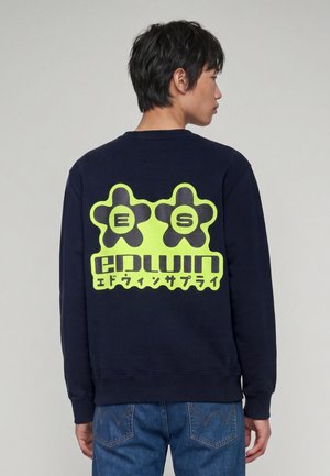Giovane uomo di profilo indossa una felpa blu navy con grafica gialla di due forme di fiori e testo sul retro, abbinata a jeans blu.