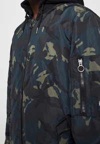 Kurtka w kamuflażu w ciemnozielonych i czarnych tonach, z zamykaną na zamek kieszenią, regulowanym kapturem oraz gładką teksturą.