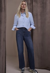 Jasnoniebieska koszula na guziki z długimi rękawami i kieszenią na piersi, w zestawieniu z ciemnoniebieskimi prostymi dżinsami i srebrnymi trampkami.