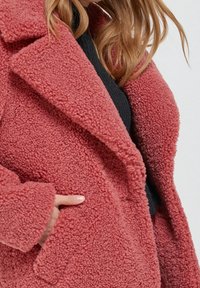 Różowy płaszcz teddy wykonany z teksturowanego, pluszowego materiału, z wyciętym kołnierzem i bocznymi kieszeniami. Charakteryzuje się luźnym krojem i przeszyciami jako detalami.