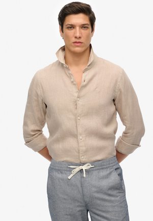 Camisa de lino beige claro con cuello abotonado y mangas remangadas, que presenta una textura sutil y un pequeño logotipo bordado.