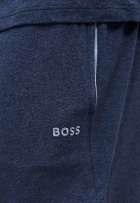 Gros plan d’un pantalon en tissu bleu foncé avec le logo brodé "BOSS" en blanc près de la poche et une bordure blanche sur le bord de la poche.