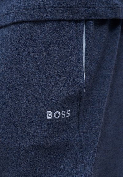 Gros plan d’un pantalon en tissu bleu foncé avec le logo brodé "BOSS" en blanc près de la poche et une bordure blanche sur le bord de la poche.