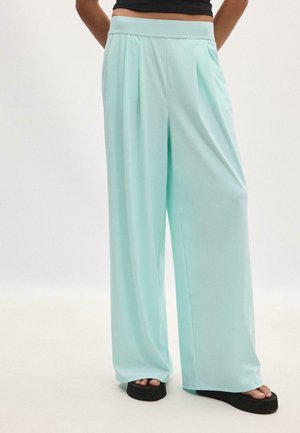 Femme portant un pantalon bleu clair à jambes larges avec des plis, associé à des sandales compensées noires et un top court noir.