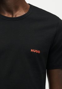 Homme portant un t-shirt noir avec un petit logo rouge "HUGO" sur le côté gauche de la poitrine.