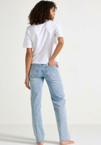 T-shirt bianco a maniche corte abbinato a jeans dritti blu chiaro, in evidenza le tasche posteriori e la vestibilità comoda. Modello in piedi a piedi nudi.