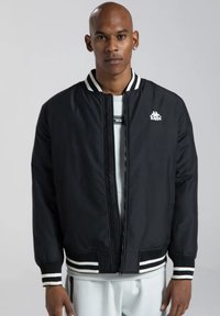 Giubbotto bomber nero con zip frontale, colletto e polsini a righe bianche, e un piccolo logo bianco sul lato sinistro del petto. Tessuto a trama liscia.