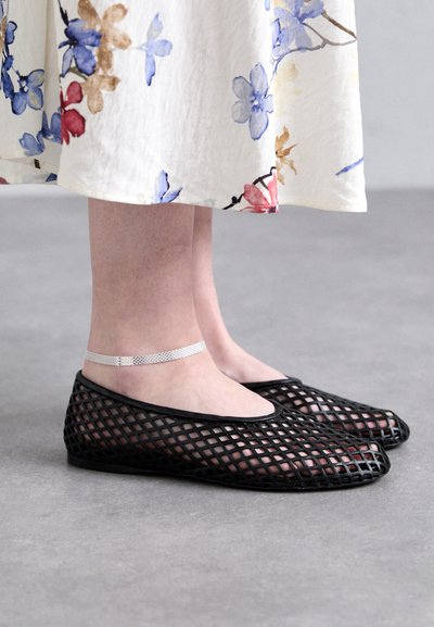 Sorte, net balletflade med et åbent, krydsdesign. Anklen prydes af et tyndt, metallic sølv bånd. En blomstret, lyse farvet nederdel hænger over.