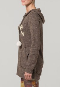 Tunic con cappuccio marrone realizzata in tessuto testurizzato, con una grande tasca e dettagli in pompon bianchi, decorata con scritte e un motivo di trifoglio.