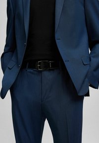 Blauw colbert met een gestructureerde afwerking, gecombineerd met bijpassende broek. Zwarte riem met een rechthoekige gesp zichtbaar op de taille.