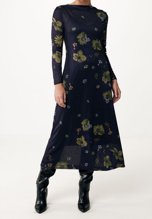 Granatowa sukienka w kwiaty z długim rękawem, z zielonymi i fioletowymi kwiatami, noszona z czarnymi, skórzanymi kozakami za kolano na jednolitym tle.