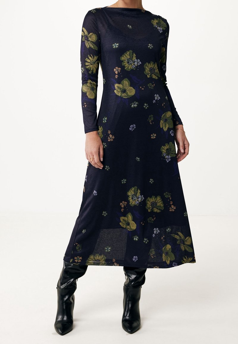 Robe fleurie bleu marine à manches longues avec des fleurs vertes et violettes, portée avec des bottes en cuir noires montant jusqu'aux genoux sur un fond uni.