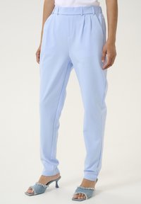 Pantaloni azzurri chiari con pieghe, una vestibilità su misura e una texture liscia. Abbinati a sandali con tacco azzurri chiari con accenti in denim sfrangiati.