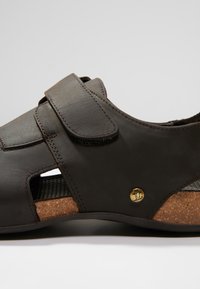 Brown leather sandal with adjustable Velcro strap, open sides for ventilation, cork sole detail, and a small gold emblem.