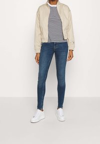 Chaqueta beige con cremallera y puños de canalé sobre una camiseta a rayas en blanco y negro, combinada con jeans ajustados azules y zapatillas blancas.