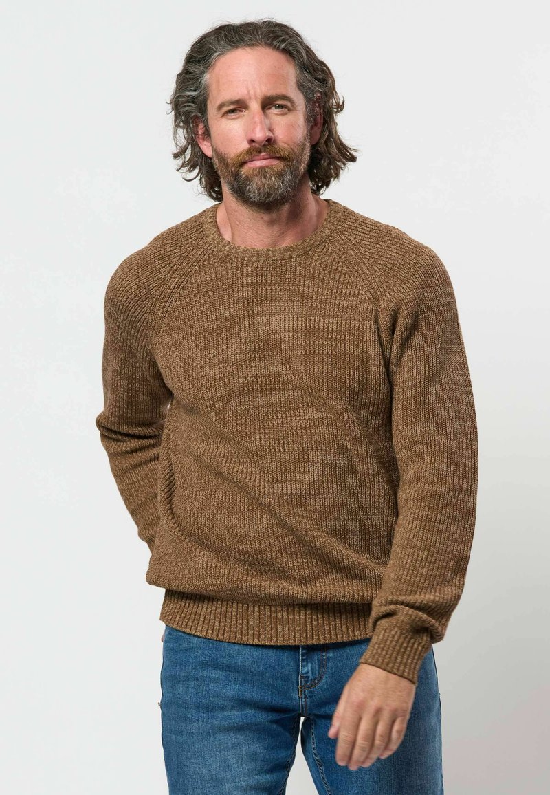 Homme aux cheveux mi-longs ondulés et à la barbe, portant un pull texturé marron et un jean bleu, debout devant un fond uni clair.