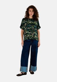 Grünes gemustertes Kurzarmoberteil kombiniert mit weit geschnittenen blauen Jeans aus Denim, die einen kontrastierenden hellen Denim-Saum und schwarze Schuhe tragen.