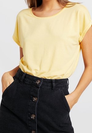 Frau trägt ein gelbes Kurzarmshirt, das in einen hoch taillierten schwarzen Jeansrock mit Knöpfen vorne gesteckt ist, mit den Händen in den Taschen.