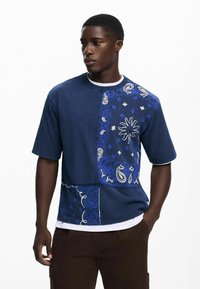 Blå denim kortærmet skjorte med et paisley-mønster på den ene side, kontrastsyninger og en rund halsudskæring, lagd over en hvid t-shirt.