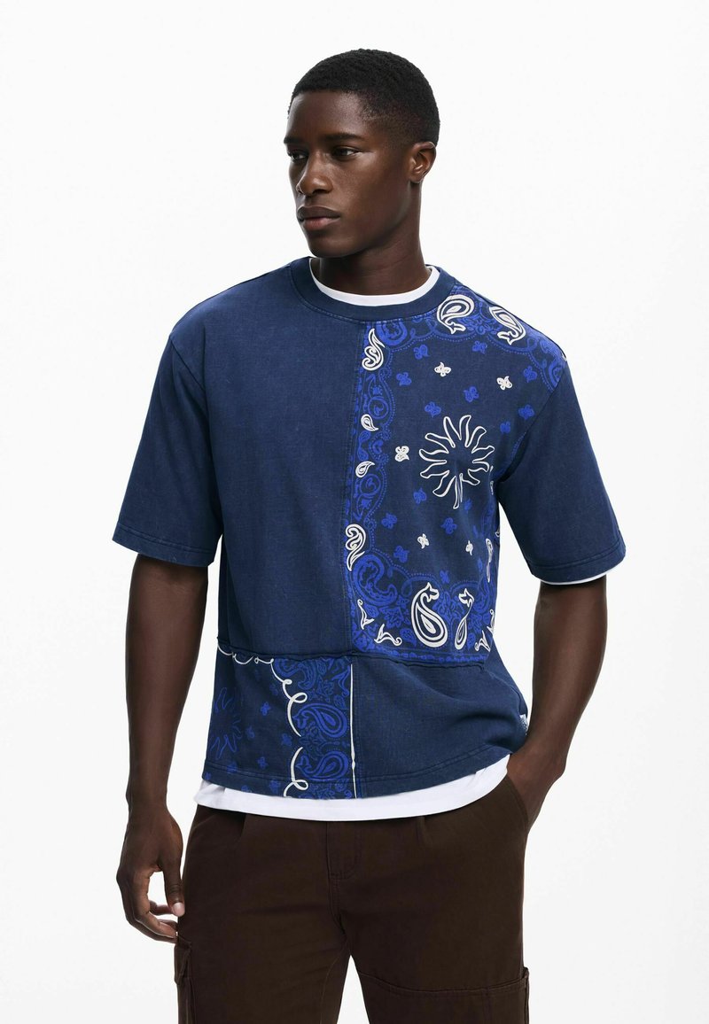 Blå denim kortærmet skjorte med et paisley-mønster på den ene side, kontrastsyninger og en rund halsudskæring, lagd over en hvid t-shirt.