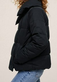 Sort pufferjakke med høj krave, der har et quiltet design og sidelommer, parret med lyseblå jeans.