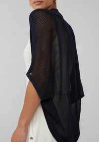 Marineblauer, gestrickter Kimono-Stil-Wickel mit offener Maschenstruktur, weiten Ärmeln und einem drapierten Design, über einem hellen Kleid getragen.