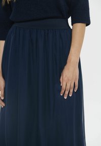 Žena u tamnoplavom pletenom puloveru i lepršavoj suknji, stoji s desnom rukom opuštenom uz tijelo na bijeloj pozadini.