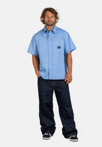 Man with curly hair wearing light blue short-sleeve button-up shirt, dark loose jeans, and black sneakers, standing with hands in pockets.