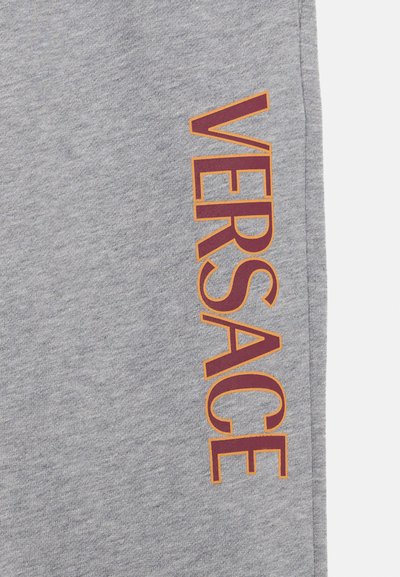 Sive bombažne trenirke z velikim logotipom "VERSACE" v burgundski in oranžni barvi ob strani. Tkanina ima mehak, teksturiran videz.