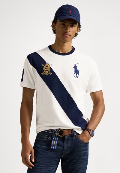 Λευκό κοντομάνικο πόλο με ναυτικό διαγώνιο ρίγα, κεντημένα λογότυπα, συνδυασμένο με σκούρο τζιν και καφέ ζώνη με ριγέ λεπτομέρεια.