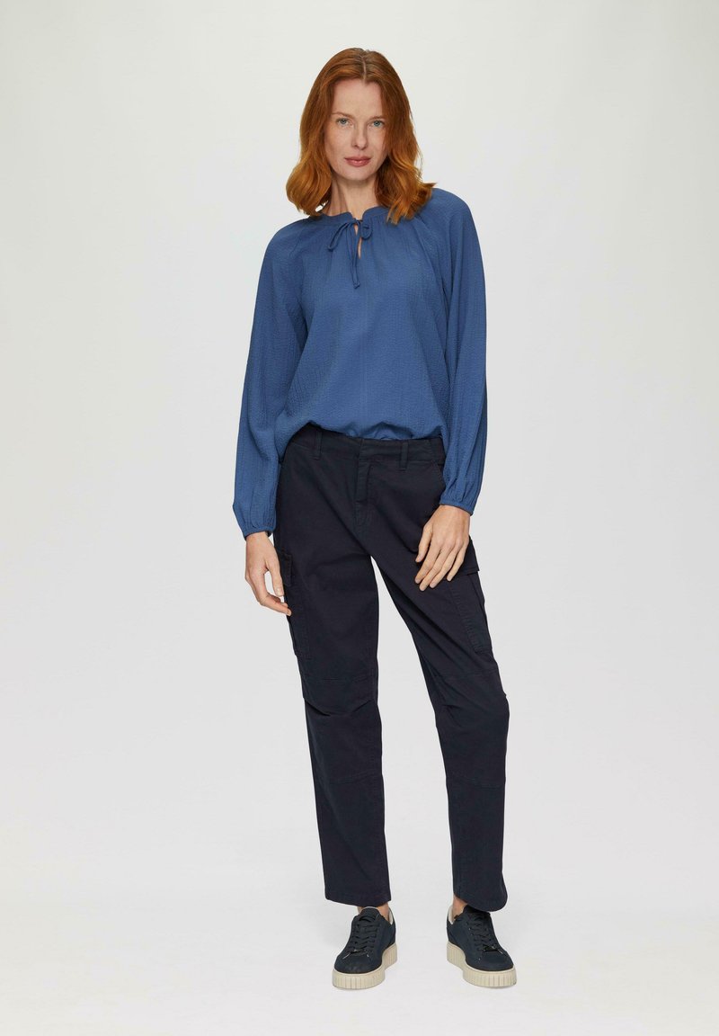 Blauwe blouse met lange mouwen en een strik bij de hals, gecombineerd met zwarte cargo-broeken. Model draagt zwarte sneakers met een gemêleerde zool.