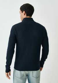 Navyblaues Strickpullover mit hohem Kragen und gerippten Bündchen, von hinten gesehen. Der Stoff erscheint weich und texturiert. Getragen mit hellen Jeans.