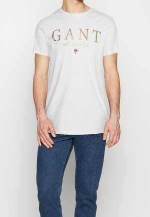 White short-sleeve t-shirt featuring gold lettering "GANT EST. 1949 U.S.A" and a small shield logo. Soft cotton material, classic crew neckline.