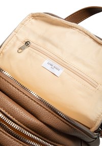 Brązowe torba skórzana o teksturowanym wnętrzu z kremową podszewką, z zamykaną kieszenią i tkaną metką z napisem "Gerry Weber". Widoczne akcenty zamka.